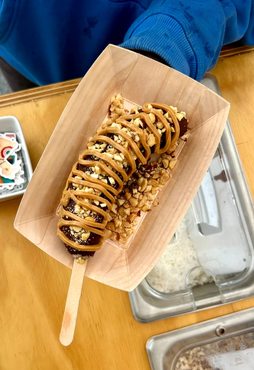A banana dessert topped with caramel drizzle, chocolate sauce, chopped nuts, and crumbled candy, served in a light wood paper tray on a wooden table.