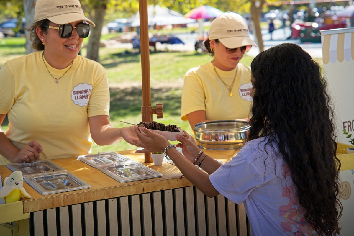 Three women in matching pale yellow t-shirts and beige caps stand behind a wooden food cart at an outdoor market, serving a customer. The shirt features a circular logo that reads 'BONNIE AND LLYAH'.