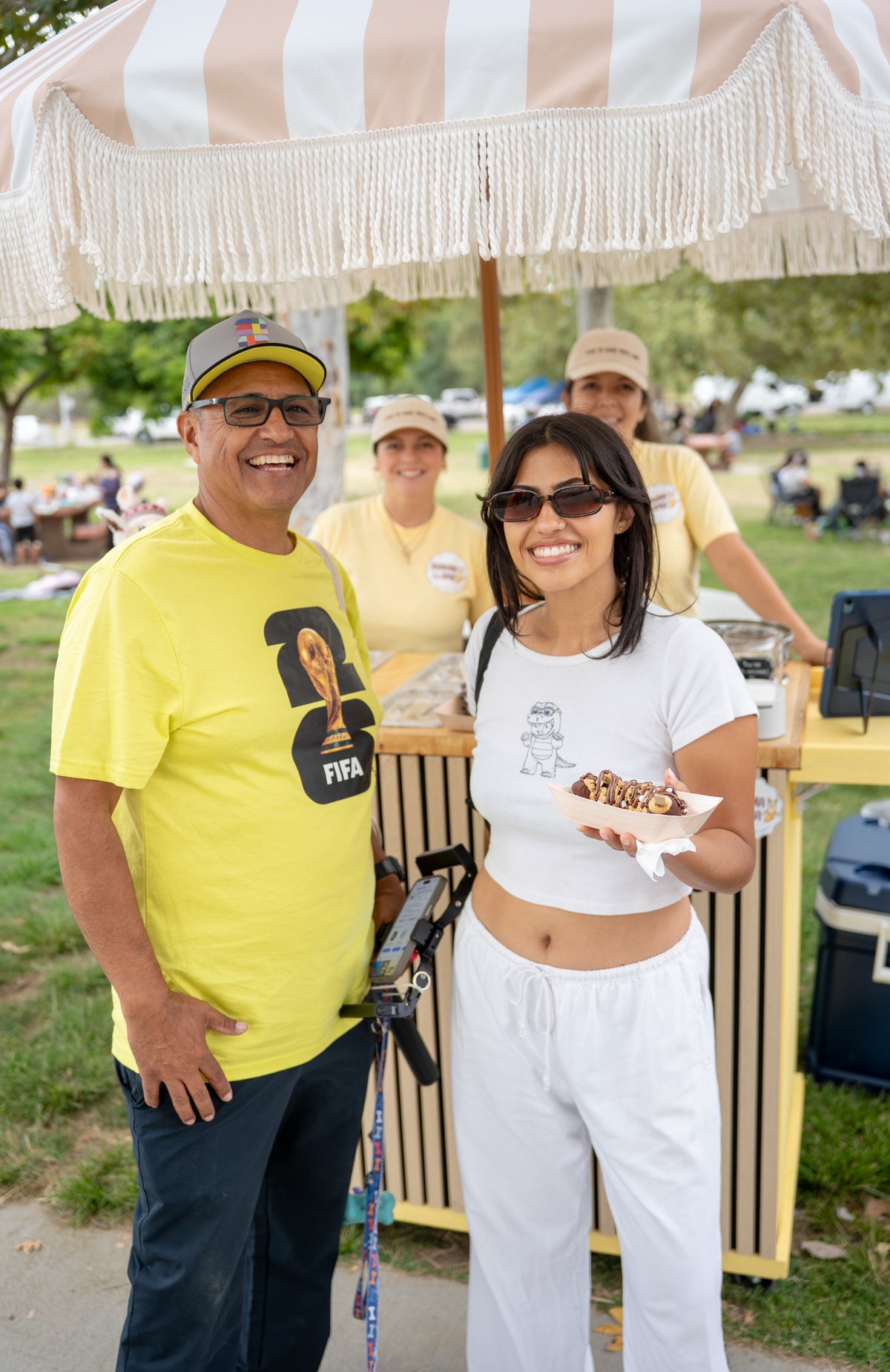 A man and a woman standing outdoors at what appears to be a fair or market booth under a beige and white striped umbrella. The man wears a bright yellow T-shirt with a black graphic and a baseball cap, while the woman wears a white crop top and white sweatpants and holds a paper tray of food.