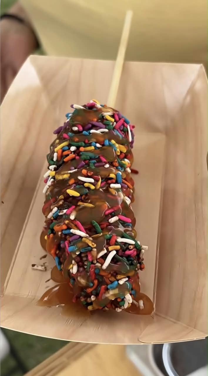A caramel-topped corn dog covered in colorful sprinkles, presented on a wooden stick inside a light brown paper tray.