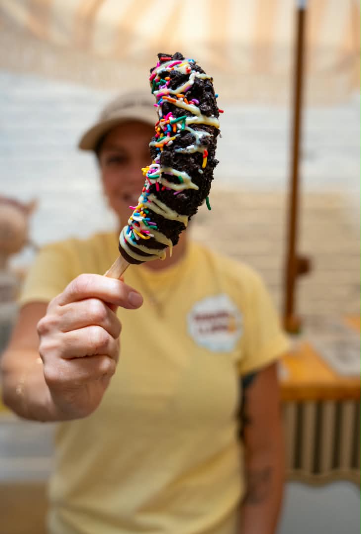 A person in a yellow shirt holds up a chocolate-covered skewer with white drizzle and colorful sprinkles, with a blurred background.