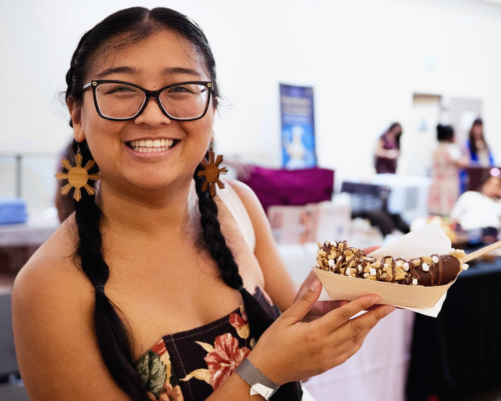 A smiling woman with braided hair and glasses holds a paper tray of chocolate-covered nuts at what appears to be an indoor event or fair. The background is softly blurred with tables and people visible.