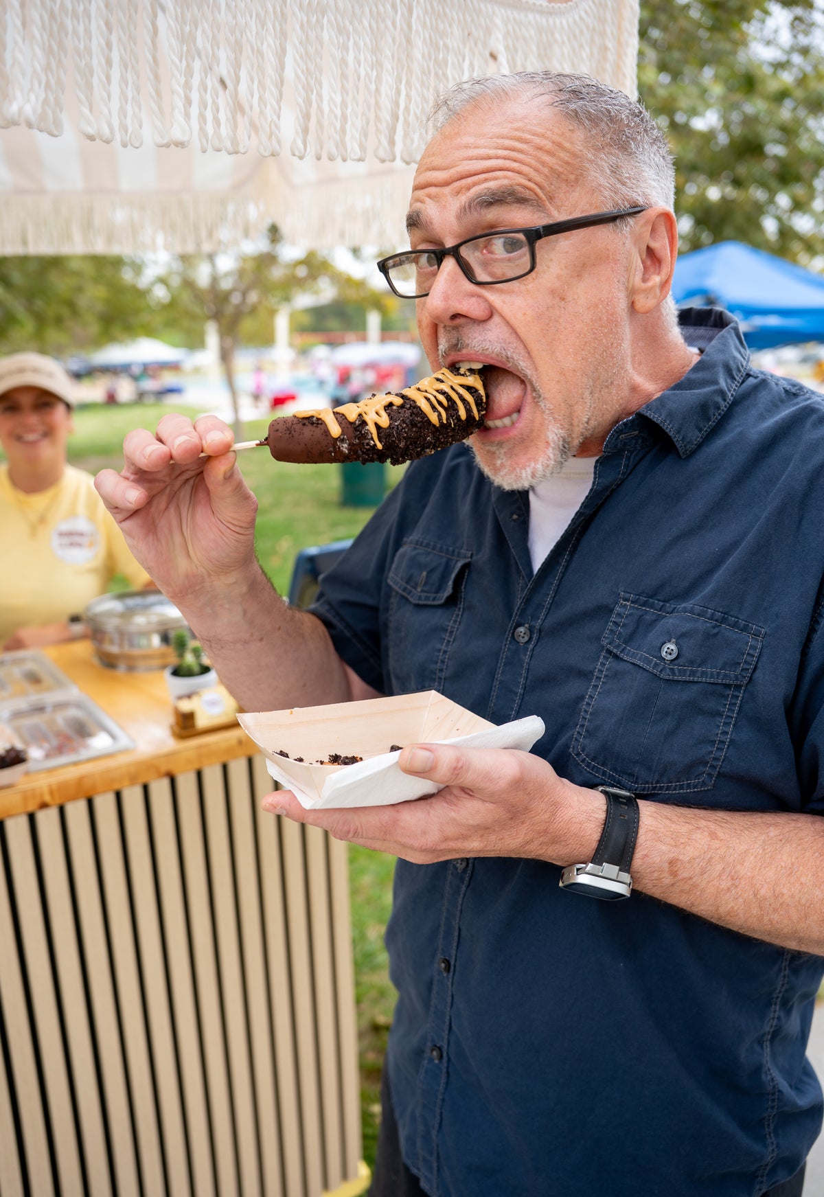 A man wearing glasses and a navy blue shirt is shown outdoors at what appears to be a market or fair, eating a chocolate-covered snack from a paper tray while holding a small dish.