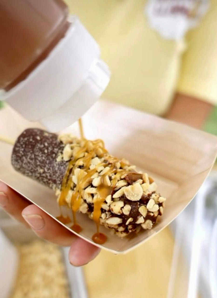 A close-up of a chocolate-covered banana on a stick, topped with chopped peanuts and drizzled with caramel sauce, being topped with more caramel from a squeeze bottle while held in a hand.