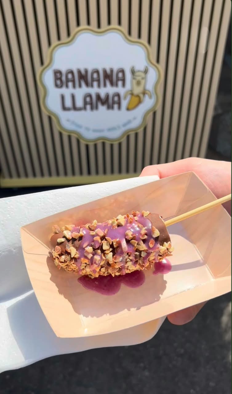 A hand holding a banana coated in chocolate and purple sauce, topped with chopped nuts, in a paper tray. The background shows a striped sign that reads 'BANANA LLAMA'.