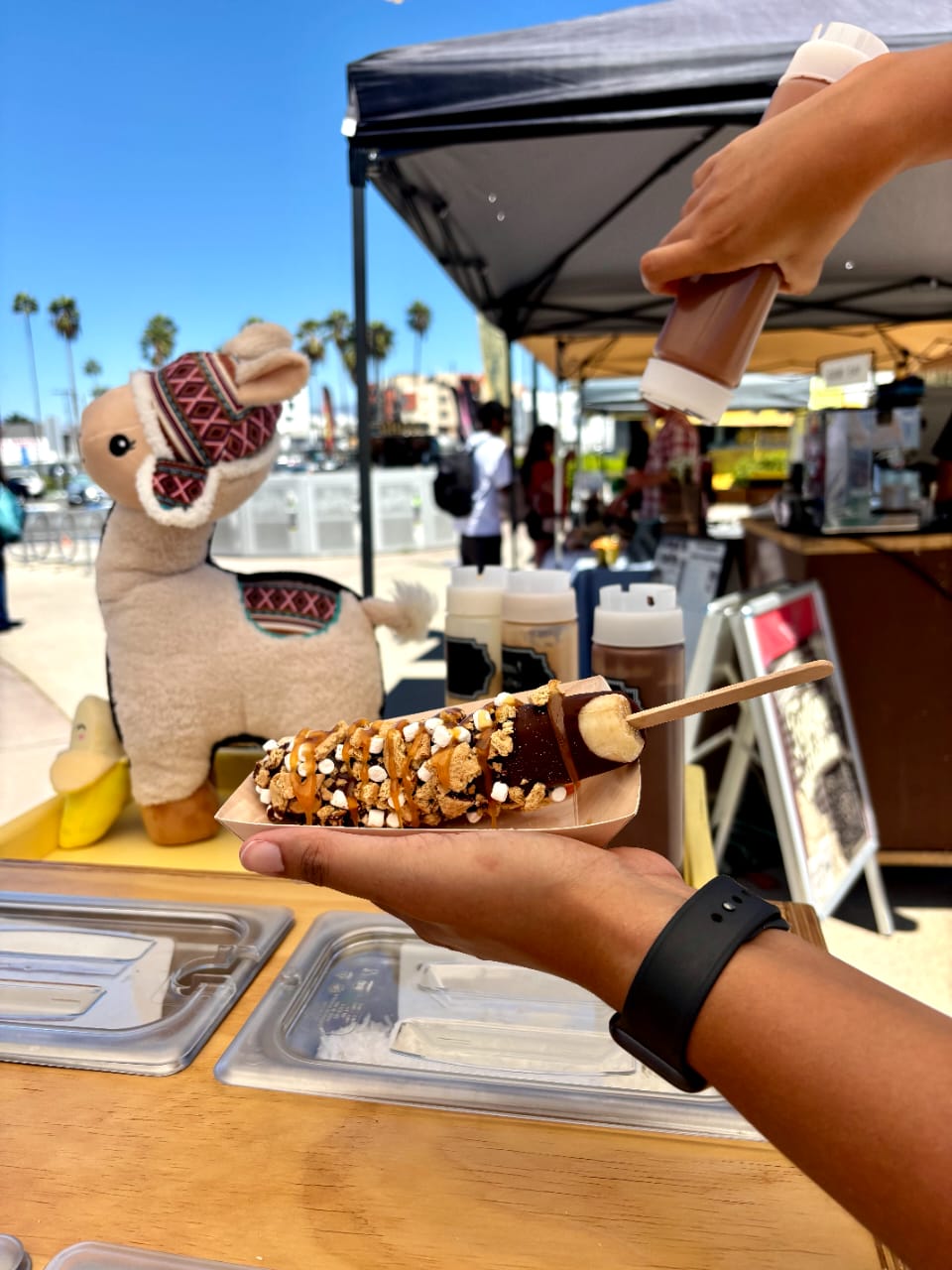 A person at an outdoor market stall is drizzling chocolate sauce onto a banana covered with nuts and marshmallows. A plush llama toy and market canopy are visible in the background under a clear blue sky.