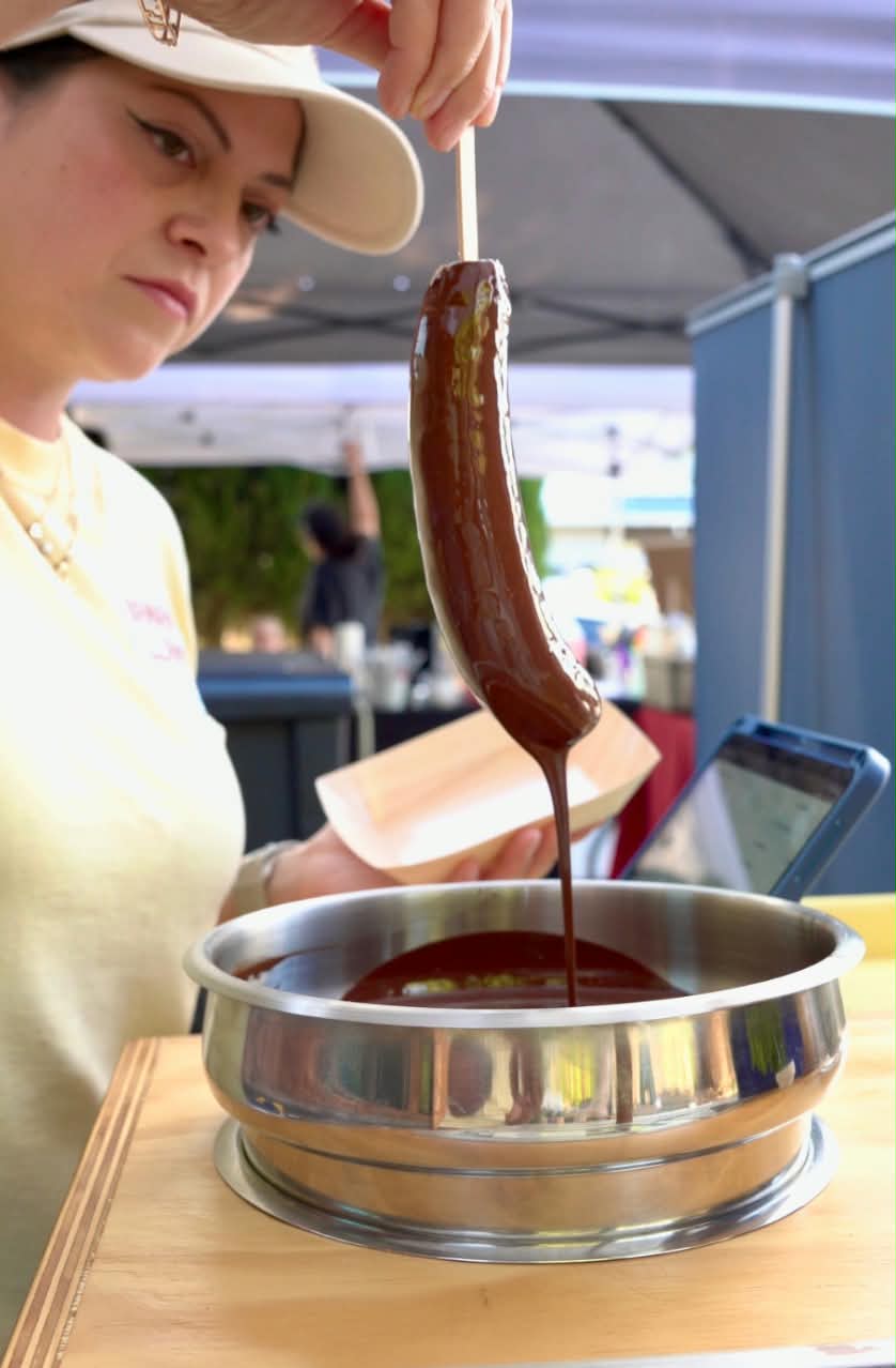 A woman at an outdoor market or fair dips a chocolate-covered banana on a wooden stick into a metal bowl of melted chocolate