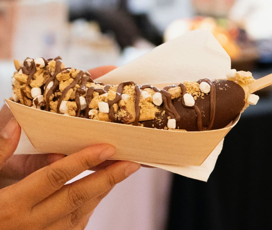 A chocolate-covered ice cream bar topped with crumbled cookie pieces and drizzles of chocolate sauce, held in a paper tray on a stick by a person's hand.