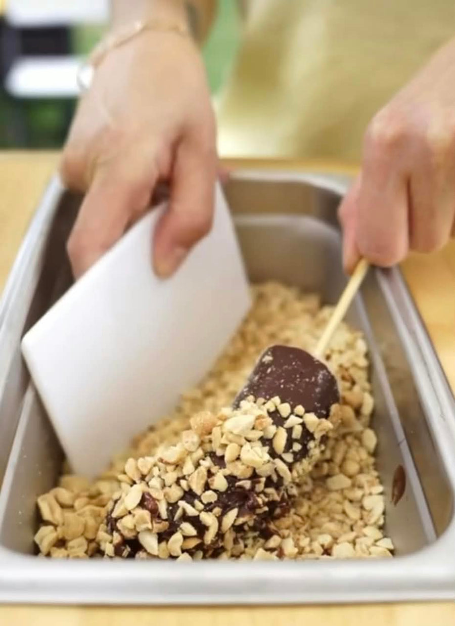 A close-up of a person's hands dipping a chocolate-covered stick into a tray filled with crushed peanuts and chocolate coating.